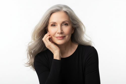 older woman with soft grey hair slightly blowing in the wind, gently smiling with her hand under her chin, wearing a black shirt, background is white