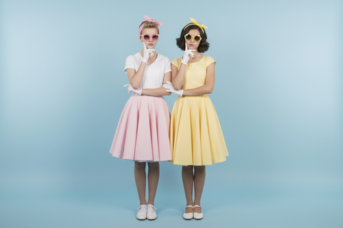two teenage girls, vintage style 50's with pink and yellow sunglasses, arms looped together, each free arm is resting on the chin with their free arms slightly bent